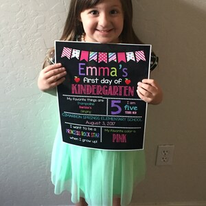 May include: A black chalkboard sign with colorful decorations announcing a girl's first day of kindergarten. The sign includes her name, age, favorite things, and school. The girl is holding the sign and smiling at the camera.