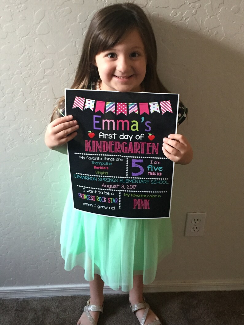 May include: A black chalkboard sign with colorful decorations announcing a girl's first day of kindergarten. The sign includes her name, age, favorite things, and school. The girl is holding the sign and smiling at the camera.
