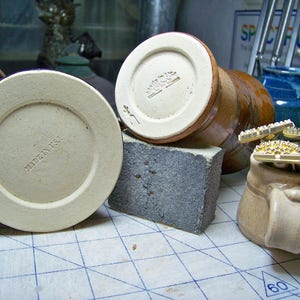 Puede incluir: Tres tazas de cerámica con diferentes diseños. El fondo de una taza dice "Made in USA". El fondo de otra taza dice "Made in USA". La tercera taza tiene un sello de metal dorado en la parte superior.