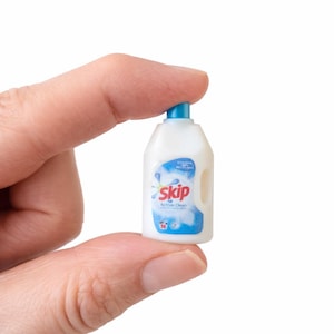 May include: A miniature white plastic bottle of Skip Active Clean laundry detergent with a blue cap and label. The label features the brand name in pink and a blue and white design. The bottle is held between fingers.