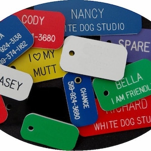 May include: An assortment of colourful rectangular dog tags with various text and phone numbers. The tags are made of plastic and have a hole for a ring. Some of the tags have a white background with black text, while others have a black background with white text. The tags are arranged in a random pattern on a black surface.