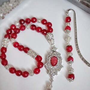 Puede incluir: Un collar de cuentas rojas con un colgante de plata que presenta una gran piedra roja rodeada de piedras transparentes más pequeñas. El collar se combina con un marcapáginas de plata con cuentas rojas.