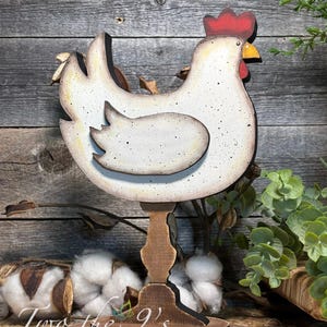May include: Wooden chicken figurine with a white body, speckled with brown spots, and a red comb. The chicken is mounted on a brown stand with a round base. The design has a rustic, farmhouse aesthetic.