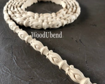 Op de afbeelding: Een close-up van een houten lijst met een decoratief patroon. De lijst is ongeverfd en heeft een natuurlijke houtkleur. De lijst is opgerold in een cirkel, met een recht gedeelte dat naar buiten steekt. De tekst "WoodUbend" is zichtbaar op de lijst.