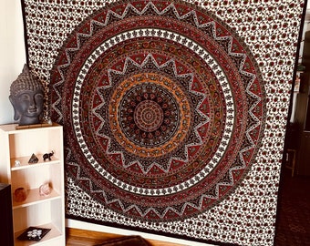 May include: A black and white tapestry with a large, colorful mandala design in the center. The mandala features intricate patterns in shades of red, orange, yellow, green, and blue. The tapestry is hanging on a wall and there are two brown cushions in the foreground.
