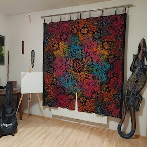 Może przedstawiać: Pokój z kolorową tapiserią wiszącą na ścianie, futerałem na gitarę na podłodze i drewnianą rzeźbą jaszczurki po prawej stronie obrazu.