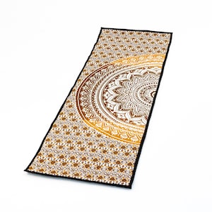 Puede incluir: Una esterilla de yoga larga y rectangular con un borde negro y un diseño de mandala dorado y blanco. El mandala es un diseño de medio círculo con detalles intrincados.