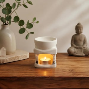 May include: A white ceramic oil burner with a square base and a bowl-shaped top. The central opening contains a small, lit tea light candle with a warm yellow flame. The burner features a smooth, matte finish and a clean, structured design.