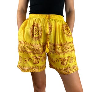 Può includere: Pantaloncini gialli con vita con coulisse e tasche. I pantaloncini presentano un motivo ripetuto di soli e il simbolo Om in marrone rossiccio. I pantaloncini sono realizzati in un tessuto leggero.