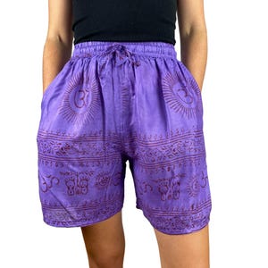 Può includere: Pantaloncini viola con vita con coulisse e tasche. I pantaloncini presentano un motivo ripetuto di simboli e disegni a forma di raggio di sole in una tonalità più scura di viola. I pantaloncini sono realizzati in tessuto leggero.