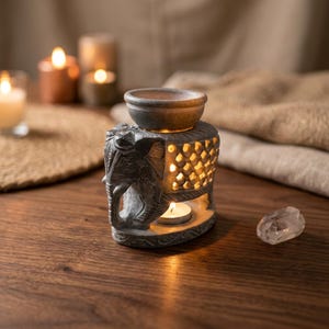 May include: A gray stone oil burner shaped like an elephant with intricate carved details and a lattice pattern on the sides. The top features a small bowl for oil, while the base contains a lit tea light candle that illuminates the interior.