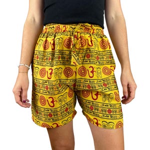 May include: Yellow shorts with a red and black pattern featuring Om symbols and other designs. The shorts have an elastic waistband with a drawstring. The shorts are made of a lightweight fabric.