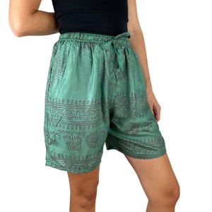Può includere: Pantaloncini verdi con vita con coulisse e un motivo ripetuto di soli e altri simboli. I pantaloncini sono realizzati in un tessuto leggero e hanno una vestibilità rilassata. I pantaloncini sono lunghi fino al ginocchio e sono adatti per l'abbigliamento casual.