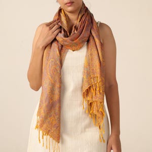 May include: A paisley patterned scarf in shades of orange, yellow, and purple. The scarf has a fringed edge and is draped over the shoulders. The scarf is made of a soft, lightweight material.