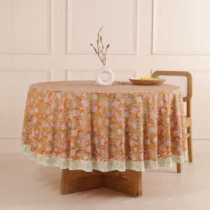 Puede incluir: Mantel redondo naranja con un estampado floral en blanco y verde. El mantel está sobre una mesa de madera. Un jarrón blanco y una silla de madera están en el fondo. El mantel tiene un borde verde.