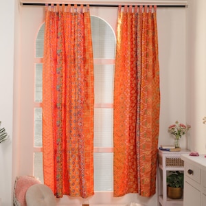 Puede incluir: Dos cortinas naranjas estampadas colgadas frente a una ventana. Las cortinas tienen un diseño de patchwork con varios patrones geométricos y florales en tonos naranja, rojo y verde. Las cortinas están colgadas de una barra negra.