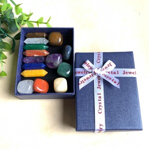 Könnte beinhalten: Eine blaue Geschenkbox mit einem weißen Band mit der Aufschrift "Crystal Jewel". Im Inneren sind eine Vielzahl farbiger Edelsteine in einer blauen Box angeordnet. Die Edelsteine umfassen klare, orangefarbene, gelbe, grüne und violette Steine.
