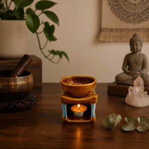 Può includere: Un bruciatore di oli essenziali in ceramica con finitura marrone lucida e accenti azzurri. Il bruciatore presenta una ciotola superiore per l'olio e una camera inferiore contenente una candela tea light accesa, che crea un bagliore caldo.
