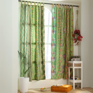 Könnte beinhalten: Grün gemusterte Vorhänge mit geometrischen und floralen Designs hängen vor einem Fenster. Die Vorhänge sind eine Mischung aus hell- und dunkelgrün mit Akzenten in Orange, Blau und Rot. Ein weißer Beistelltisch mit einer gewebten Schublade und eine Topfpflanze sind sichtbar.
