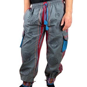 May include: Gray cargo pants with a drawstring waist, featuring black sun and paisley designs. The pants have red and white stripes down the sides, blue pocket accents, and patterned cuffs. The pants are made of a soft, comfortable material.