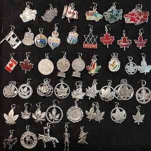 May include: A collection of silver charm pendants featuring Canadian themes. The charms include maple leaves, Canadian flags, and various city and province names. Some charms feature colorful enamel details.