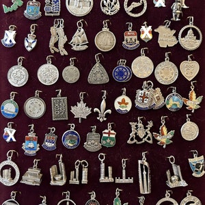 May include: A collection of silver charms featuring various Canadian city and province designs. The charms are arranged in rows on a dark red velvet background. Some of the charms feature the city name, while others feature a symbol or emblem. The charms are all in a silver finish and have a variety of shapes and sizes.