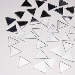 May include: A collection of small, mirrored glass triangles scattered across a white surface. The triangles reflect light, creating a shimmering effect. Some triangles appear darker, while others are lighter, adding depth to the composition. Ideal for craft projects or decorative purposes.