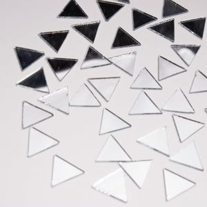May include: A collection of small, mirrored glass triangles scattered across a white surface. The triangles reflect light, creating a shimmering effect. Some triangles appear darker, while others are lighter, adding depth to the composition. Ideal for craft projects or decorative purposes.