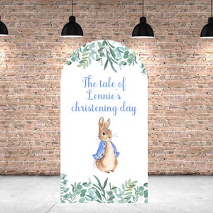 May include: A white backdrop with a green leafy border. The text "The tale of Lennie's christening day" is printed in blue. A watercolour illustration of Peter Rabbit in a blue jacket is in the centre.