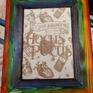 May include: A wooden picture frame with a rainbow-colored finish. The frame contains a print with a Halloween theme, featuring the words "It's just a bunch of Hocus Pocus" and images of skulls, hearts, and potion bottles.