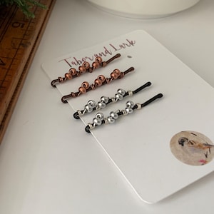May include: Set of six hair clips with copper and silver beads. The clips are black and the beads are arranged in a pattern. The card says "Tabor and Lark".