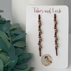May include: Two copper wire hair clips with gold and green beads. The clips are displayed on a white card with the text "Tabor and Lark" printed on it.