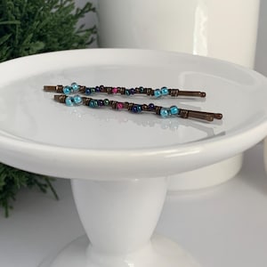 May include: Two beaded hair pins with copper-colored metal bases. The beads are a mix of blue, purple, and pink. The hair accessories are displayed on a white, pedestal-style plate. The background includes greenery and white ceramic decor.