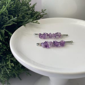 May include: Two silver hair clips with amethyst gemstone chips. The clips are on a white surface.