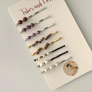 May include: A collection of beaded hair pins in various colors, including burgundy, purple, gold, and white. The pins are arranged on a white card with the text "Tabor and Lark". Each pin features a different bead design.