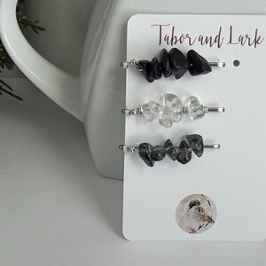 May include: Three silver-toned metal hair pins attached to a white display card labeled Tabor and Lark. Each pin features a cluster of raw, irregular stone chips in black, clear, and dark gray. A small circular sticker with a bird illustration is present.
