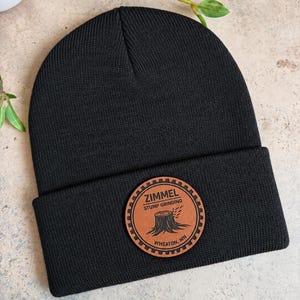 May include: A solid black knit beanie featuring a folded cuff and a circular tan leather patch centered on the front. The patch displays a gear-shaped border with the text ZIMMEL STUMP GRINDING WHEATON, MN and an illustration of a tree stump.