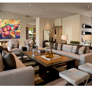 May include: A modern living room with a large, colorful abstract painting of fruit hanging above a beige sofa. The sofa is positioned in front of a coffee table with a tray of decorative items. Two beige armchairs are placed on either side of the sofa.