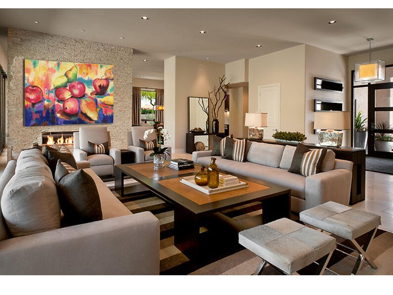 May include: A modern living room with a large, colorful abstract painting of fruit hanging above a beige sofa. The sofa is positioned in front of a coffee table with a tray of decorative items. Two beige armchairs are placed on either side of the sofa.