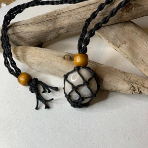 May include: A necklace featuring a light-colored stone encased in a black woven net. The necklace has a black braided cord with wooden beads. The stone is a light beige color. The necklace is displayed on a white surface with driftwood in the background.