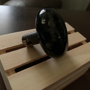 May include: A black, oval-shaped cabinet knob with a metal stem, resting on a small wooden pallet. The knob has a glossy finish with some white spots. The pallet is made of light-colored wood.