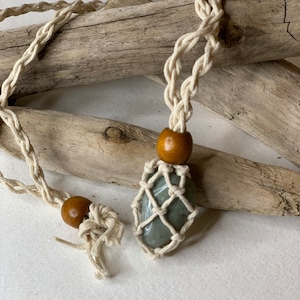 May include: A handmade macrame necklace featuring a light green stone encased in a woven net. The necklace has a natural, off-white cord with wooden beads. The design is simple and earthy, perfect for everyday wear.