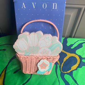 Puede incluir: Una cesta de mimbre rosa con un diseño floral y una flor rosa en la parte delantera. La cesta está llena de papel de notas blanco y rosa. La caja dice "Spring Notes Wicker Basket With 150 Sheets Of Notepaper" y "AVON".