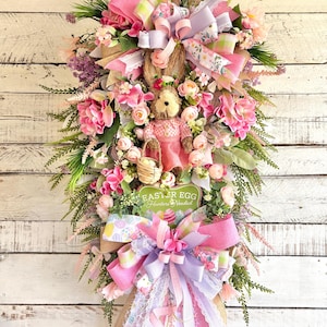 May include: An Easter-themed wreath with a plush bunny wearing a pink dress, surrounded by pink and white flowers, ribbons, and greenery. The wreath includes a sign that reads "Easter Egg Hunters Needed."