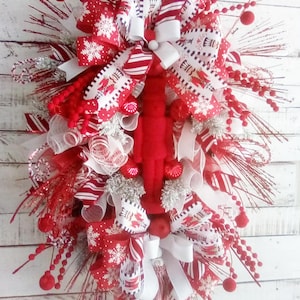 May include: A red and white Christmas wreath with a nutcracker figurine in the center. The wreath is decorated with red and white striped ribbon, red and white polka dot ribbon, and white mesh. The wreath is also decorated with red berries and silver glitter.
