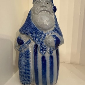 Puede incluir: Figura de cerámica de Papá Noel en tonos azules y grises. La figura lleva un gorro azul, una túnica y pantalones a rayas. La barba y la cara son grises. El artículo es un objeto decorativo.