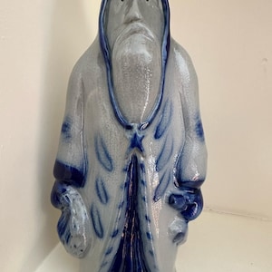 Puede incluir: Figura de cerámica de una persona con túnica, barba larga y capucha. La figura es principalmente gris con detalles azules en la capucha, el borde de la túnica y otros detalles. La figura sostiene una pequeña bolsa.