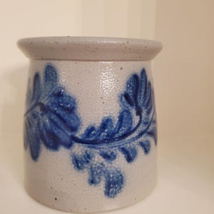 Puede incluir: Un tarro de gres con un acabado moteado gris claro y un diseño floral azul. El tarro tiene un cuerpo redondeado y un borde ligeramente ensanchado. El diseño azul presenta hojas y vides estilizadas.