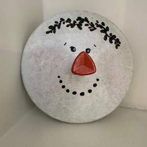 May include: A circular, white snowman face with a red carrot nose and a dotted black smile. The snowman has black dot eyes and a green and red holly berry wreath. A festive holiday decoration.