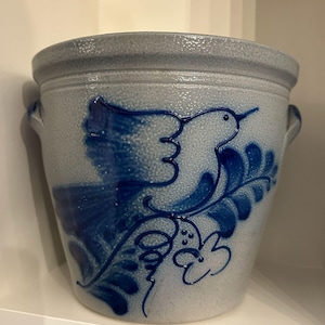 Puede incluir: Un tarro de gres con un diseño de pájaro y flores en azul y blanco. La olla de cerámica tiene dos asas pequeñas y un borde ancho. El pájaro está posado en una rama frondosa. El tarro se utiliza probablemente para guardar o decorar.